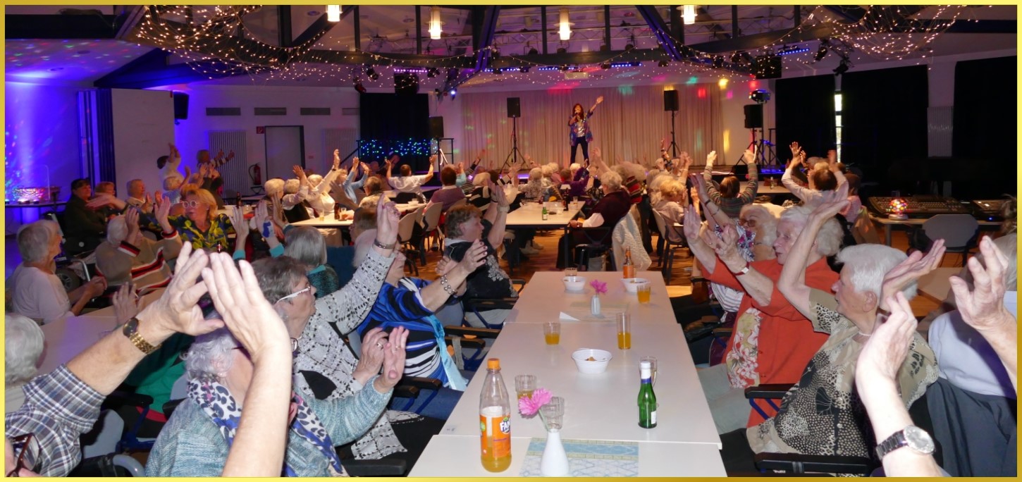 Großer Saal voller Senioren, die die Hände zum Himmel strecken.